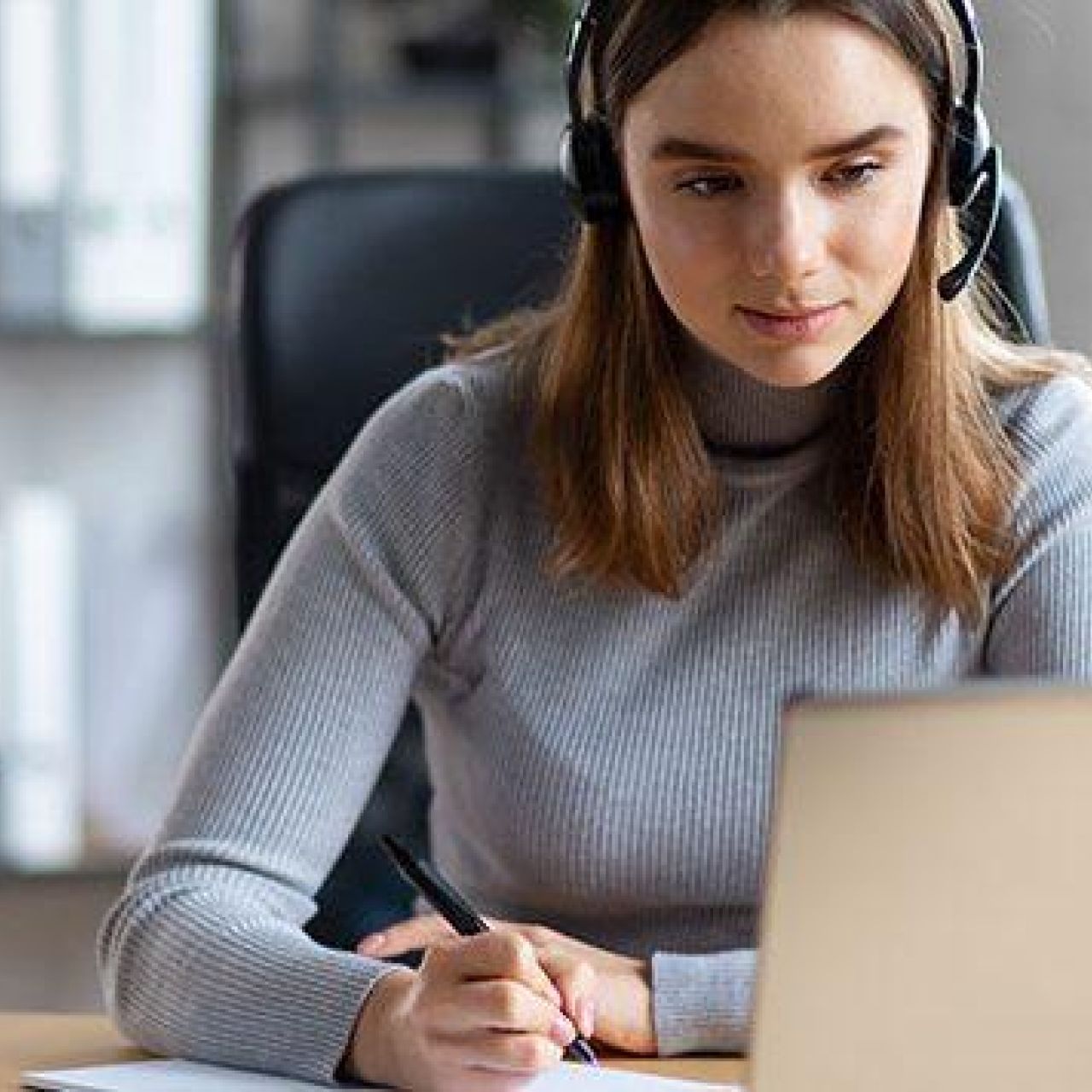 Adult wearing headphones takes notes on paper while looking at a laptop.